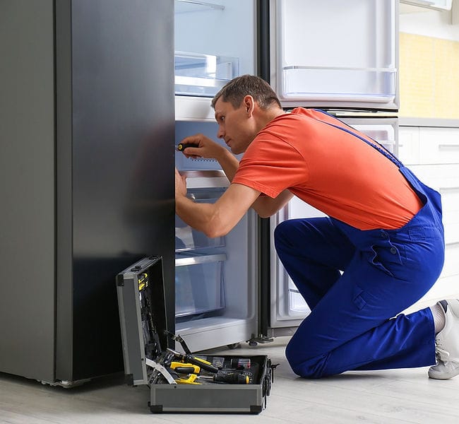 تکنسین با لباس کار در حال تعمیر یخچال ساید بای ساید در آشپزخانه، همراه با جعبه ابزار باز